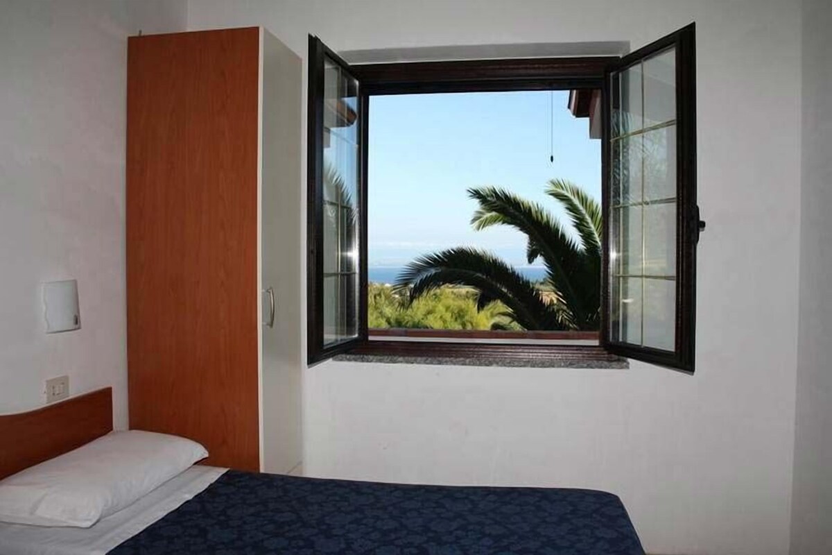 Un dormitorio con cama, armario de madera y ventana con vista a palmeras y al mar.