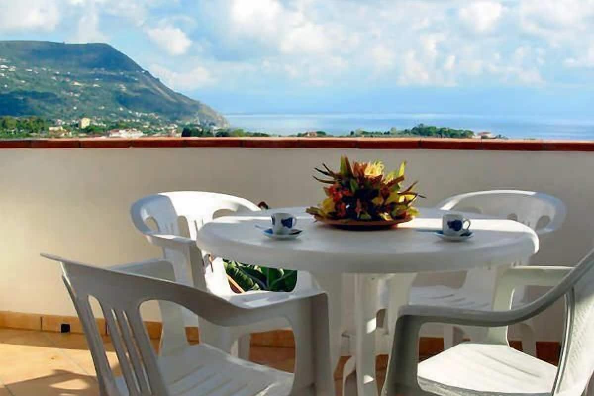 A round table with white chairs and a floral centerpiece on a terrace with a view of the sea and a mountain.