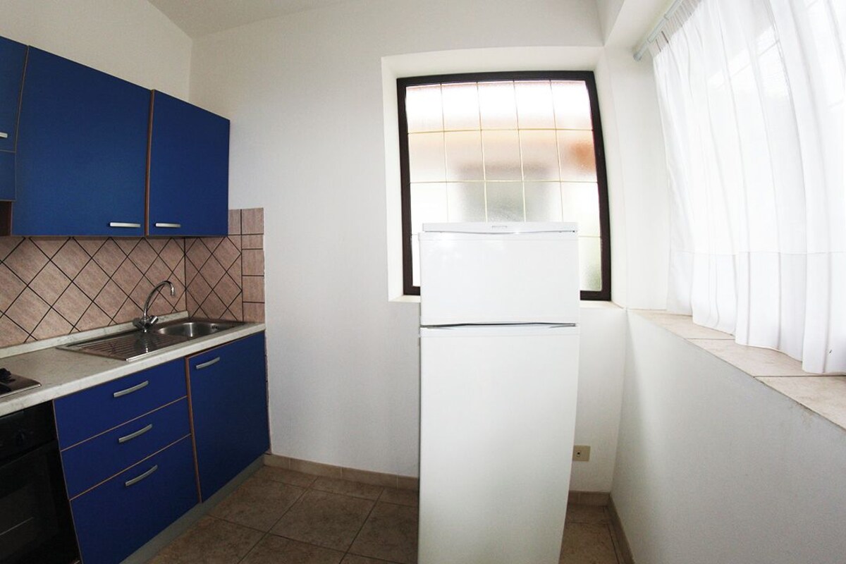 Una cocina compacta con armarios azules, un refrigerador blanco, un ventanal con vidrio esmerilado y piso de baldosas.
