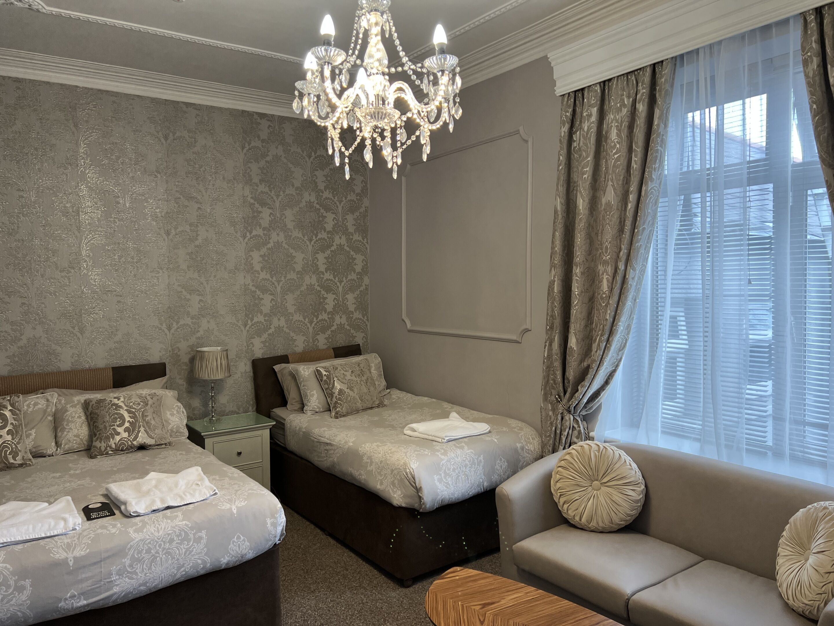 A hotel room with two beds, a sofa, a chandelier, and a large window with curtains.