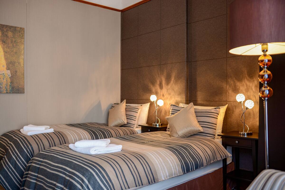 Habitación de hotel con dos camas, ropa de cama a rayas, una mesita de noche con lámpara y un cuadro en la pared.