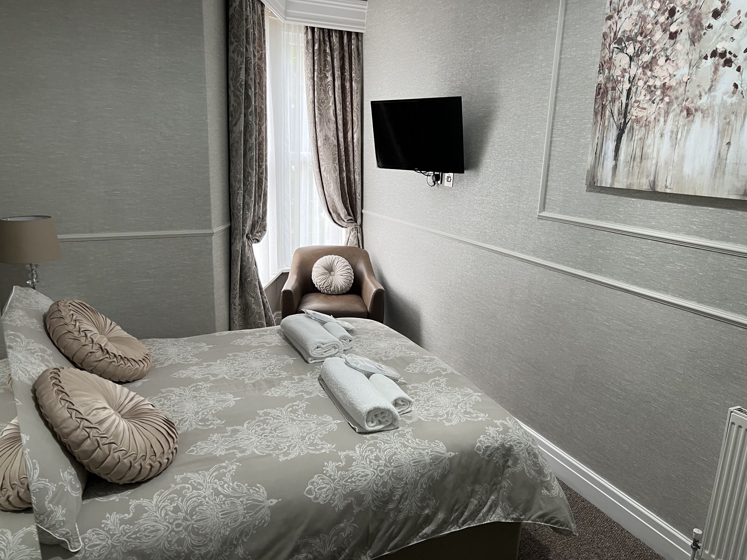 A neatly arranged bedroom with a bed, pillows, a chair, a TV, and a painting on the wall.