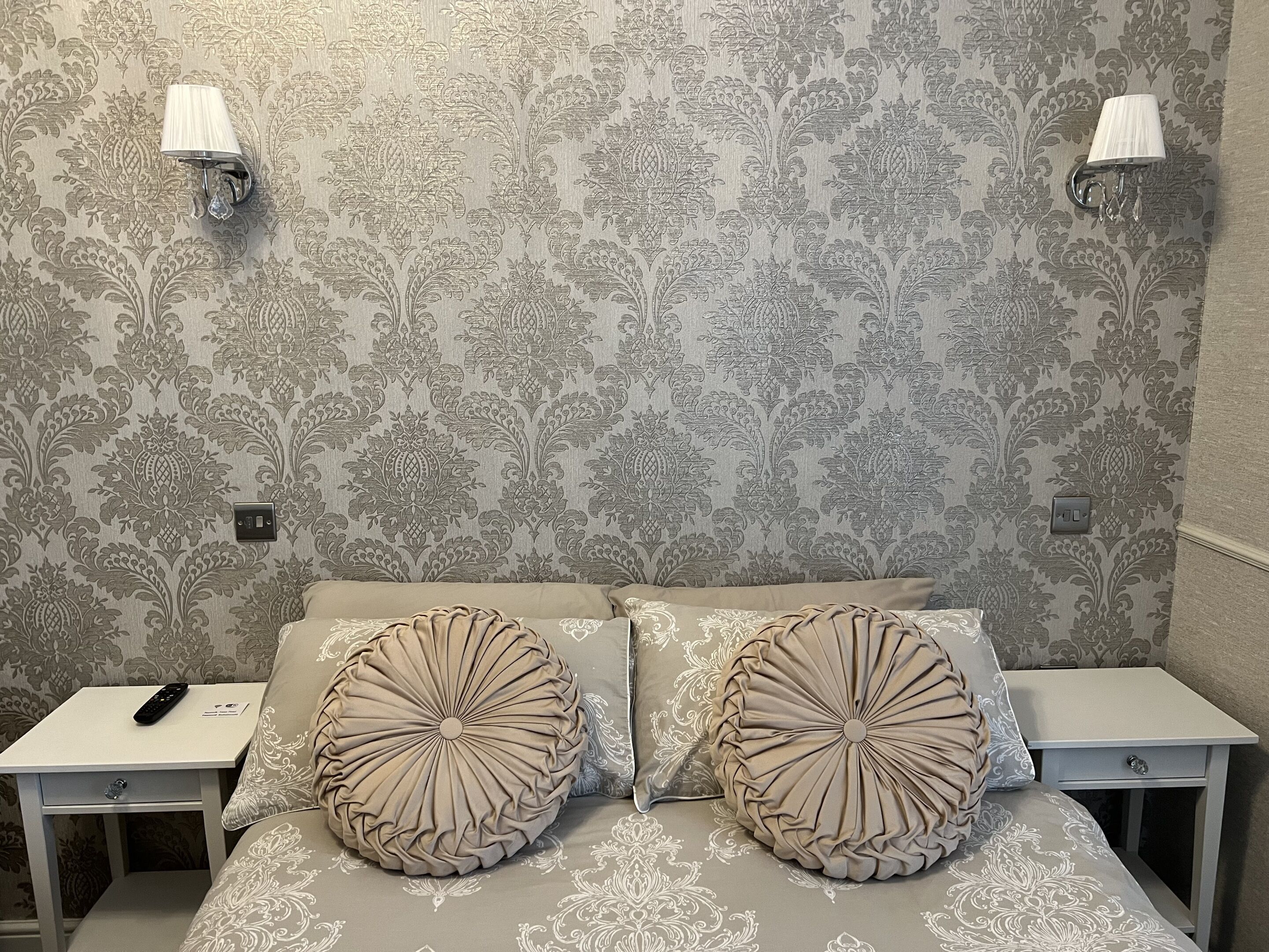 A bedroom with a patterned wallpaper, two bedside lamps, a bed with a decorative headboard, and a nightstand with a remote control.