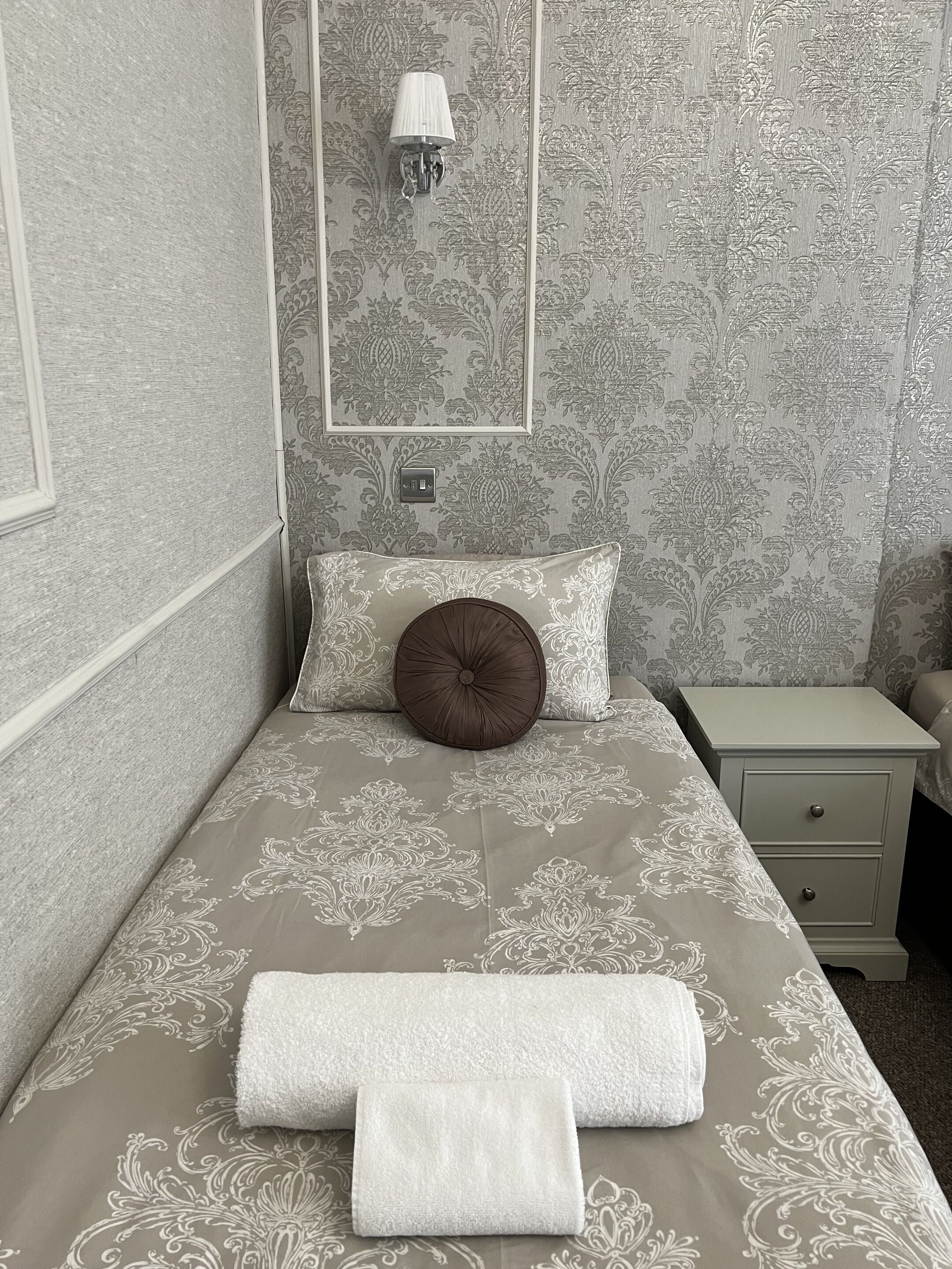 A neatly made bed with a patterned bedspread, a round brown pillow, and two folded towels.