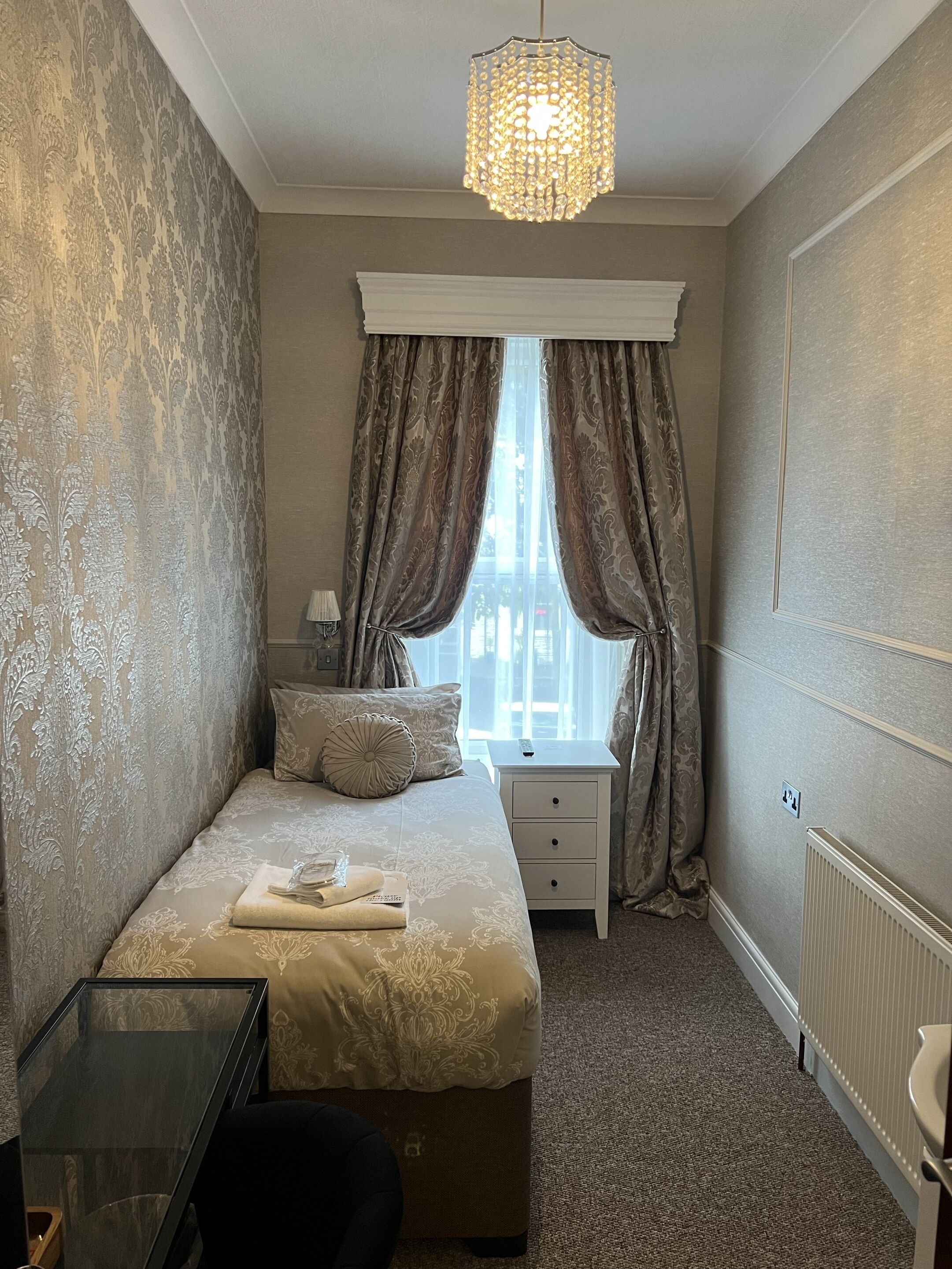 Une chambre à coucher avec un lit, une table de chevet, une lampe, une fenêtre avec des rideaux et un lustre.