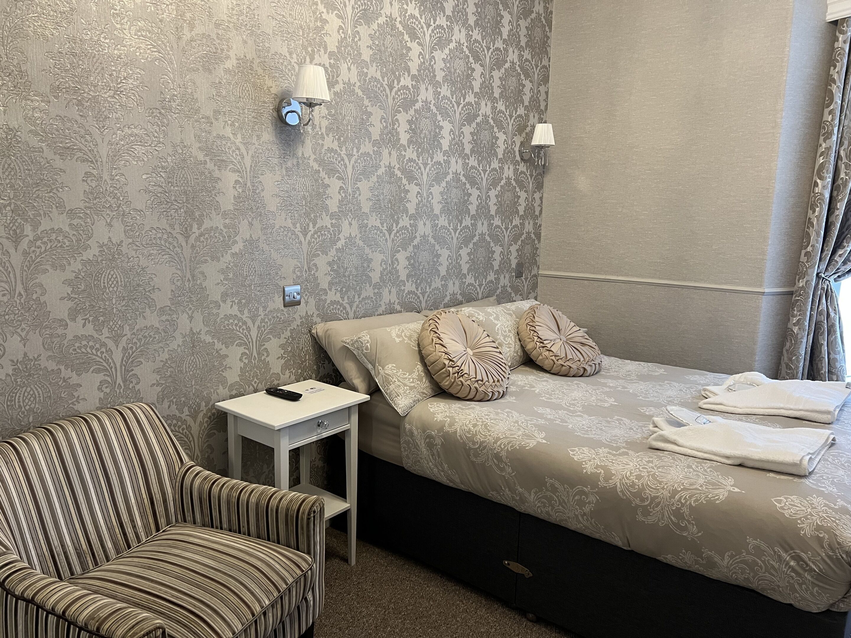 A bedroom with a bed, a striped armchair, a bedside table with a remote control, and patterned wallpaper.