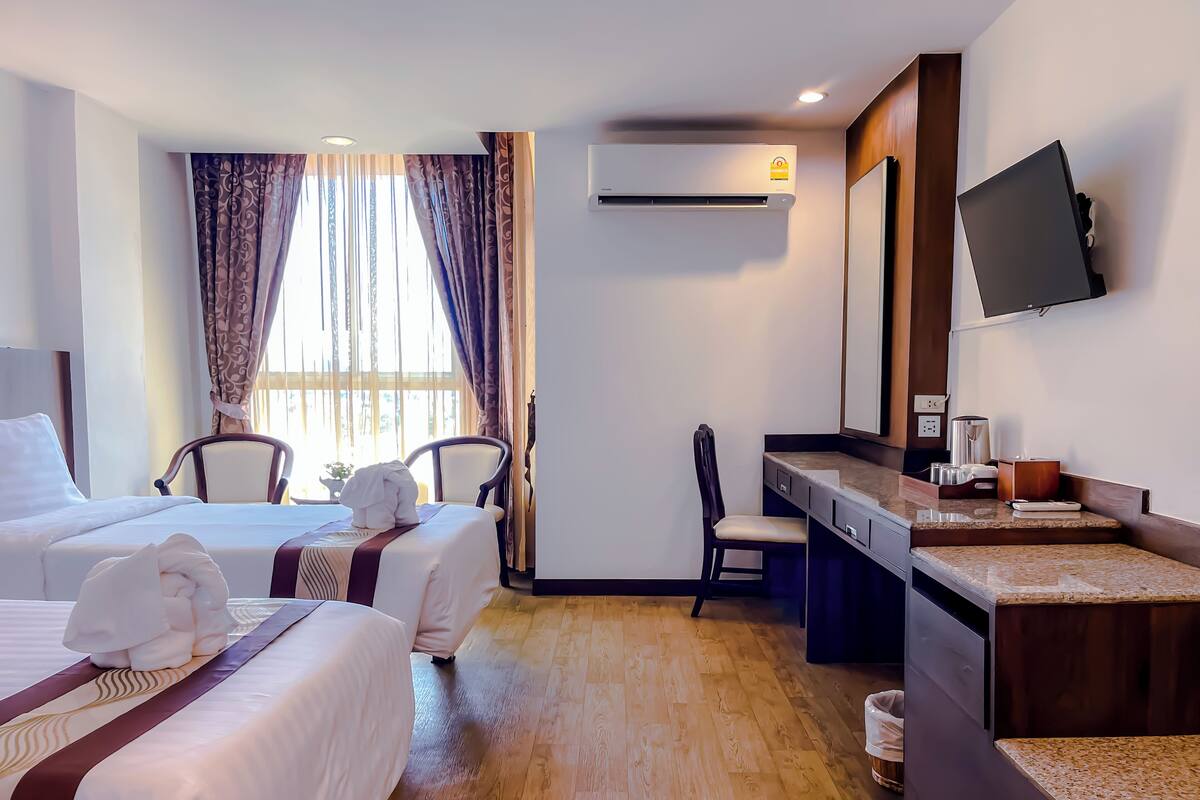 A hotel room with two beds, a desk, a chair, a TV, and an air conditioning unit.