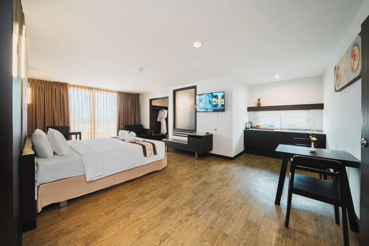 A modern hotel room with a large bed, a desk, a TV, and a kitchenette.