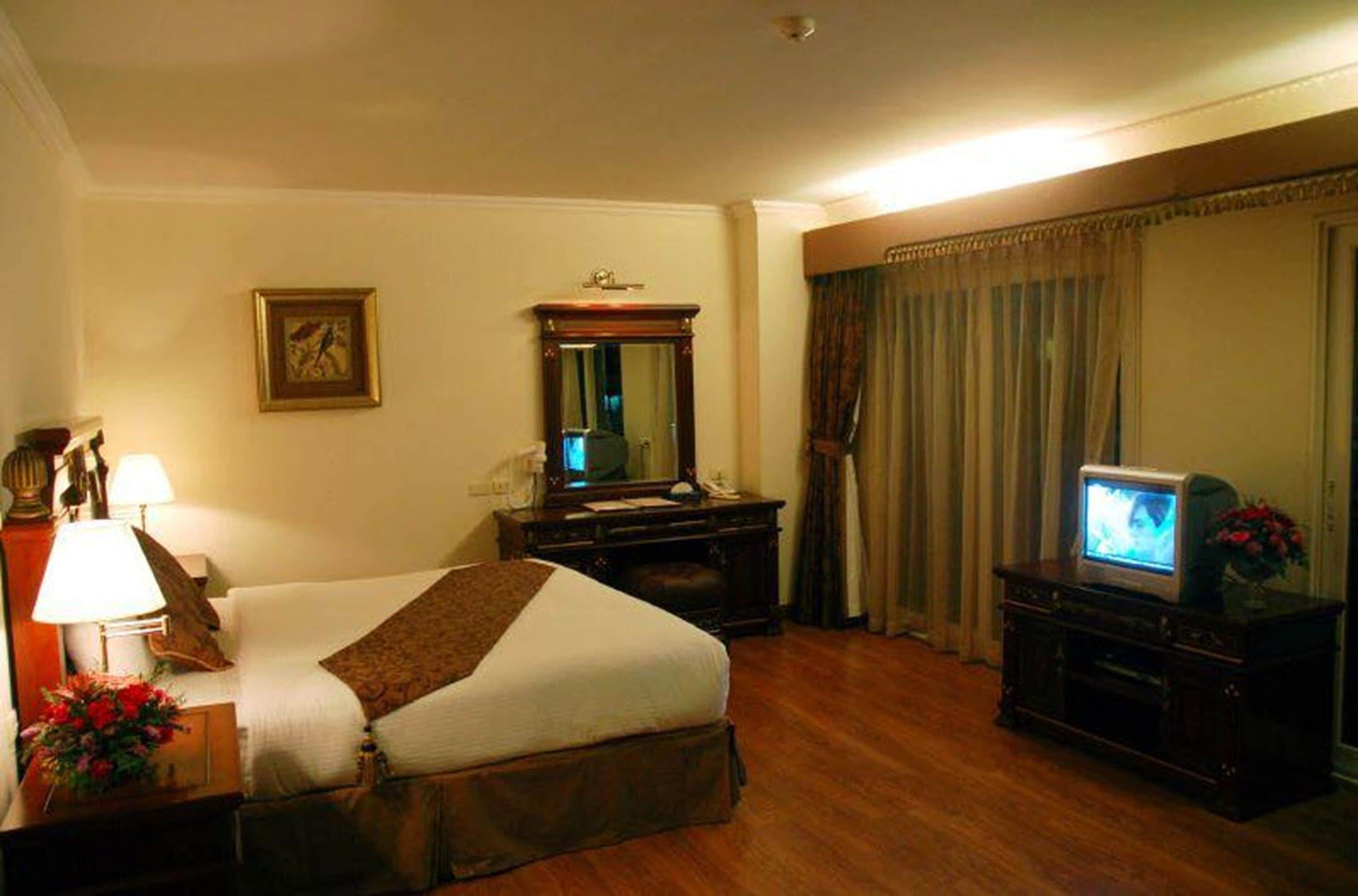 Een hotelkamer met een bed, een televisie, een kaptafel, een spiegel, een lamp en een lijst met een afbeelding aan de muur.