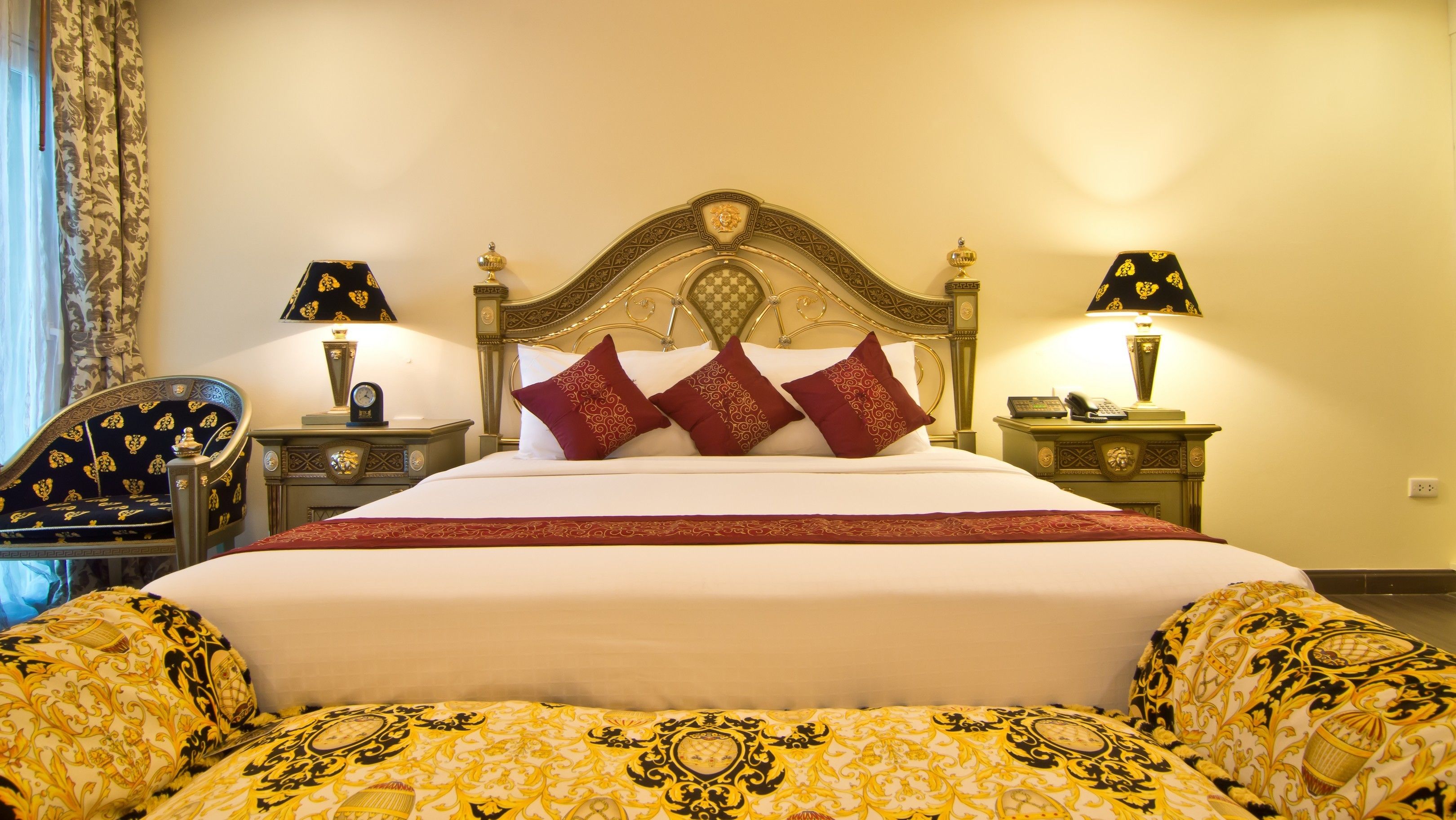 Een net opgemaakt bed met een wit-rode sprei, omringd door bijpassende kussens. Er zijn twee nachtkastjes met lampen, een stoel met een gestreept kussen en een decoratief hoofdbord.