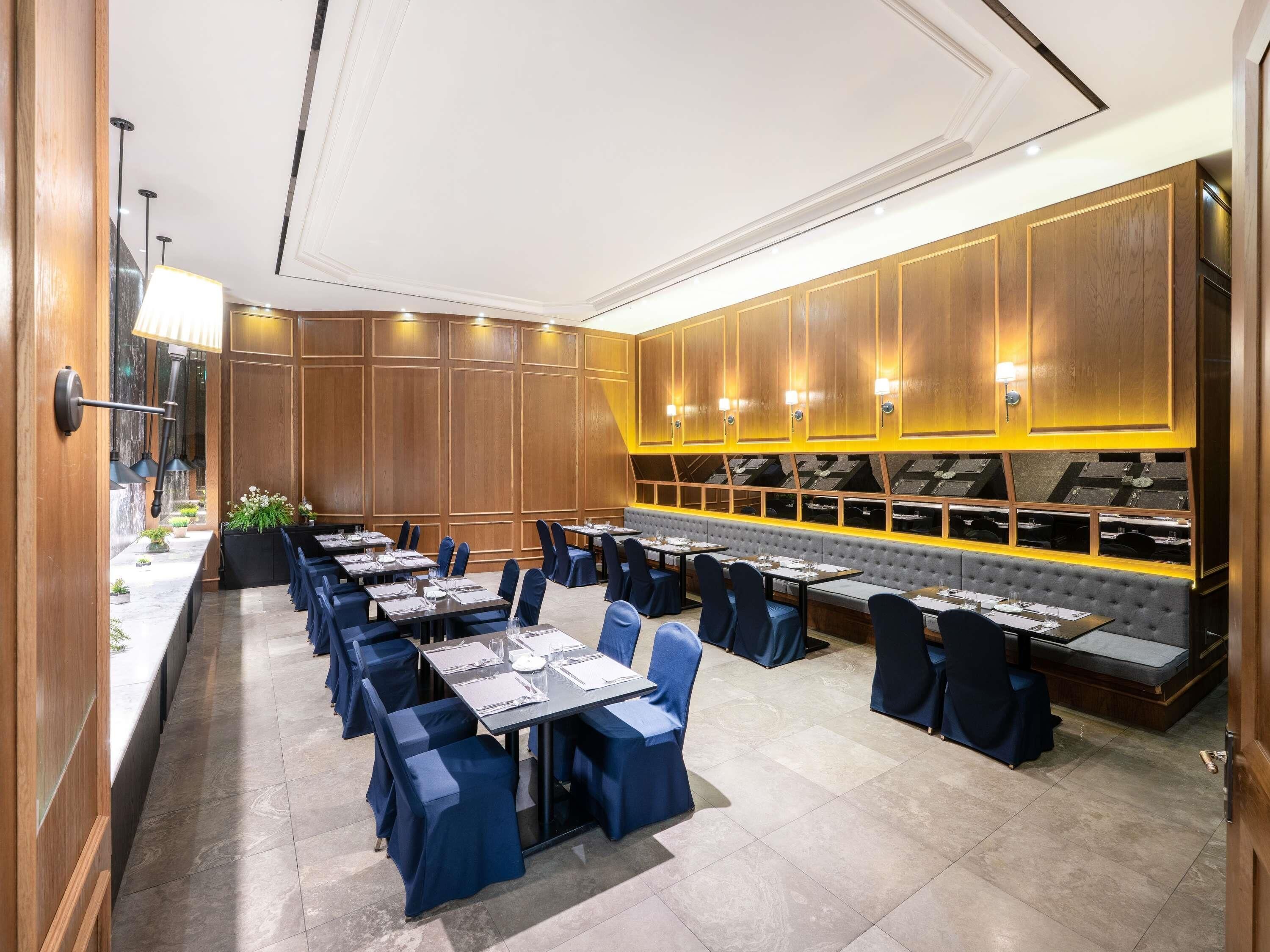 Un comedor de hotel con varias mesas y sillas, una larga barra con lámpara, y un rincón de estar con un banco acolchado.