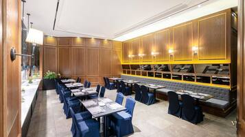 Un comedor de hotel con varias mesas y sillas, una larga barra con lámpara, y un rincón de estar con un banco acolchado.