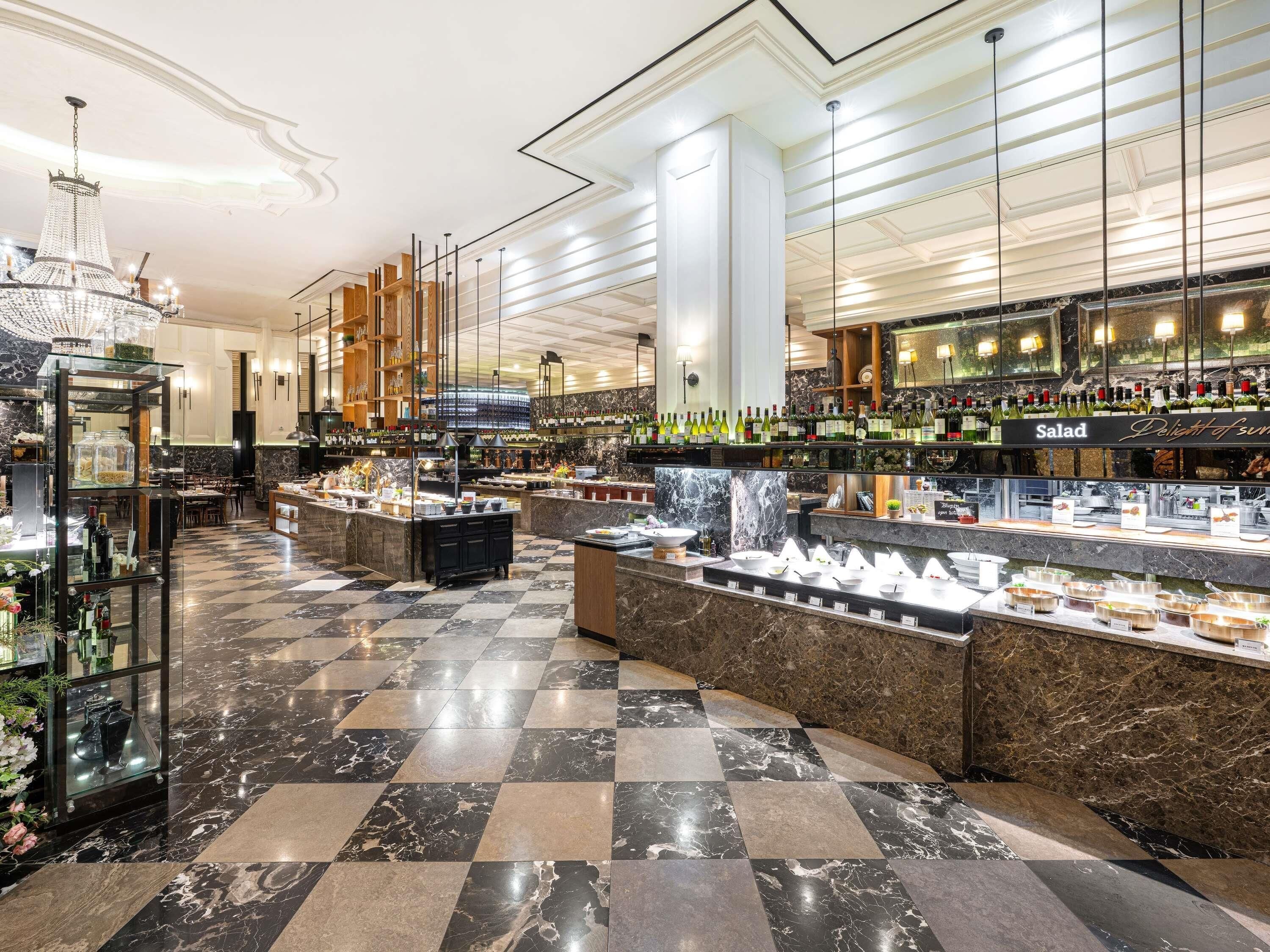 Un vestíbulo de hotel con piso de mármol a cuadros, una zona central de bar con diversos tipos de botellas y un mostrador de buffet con exhibiciones de comida.