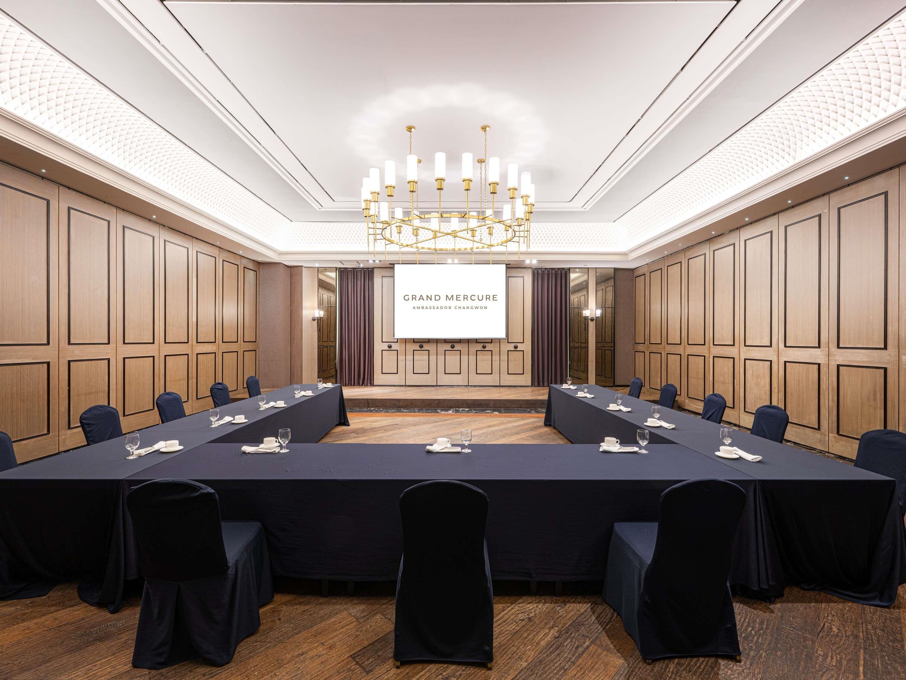 A conference room with a long table, chairs, a chandelier, and a screen displaying 'Grand Mercure Ambassador Changwon'.