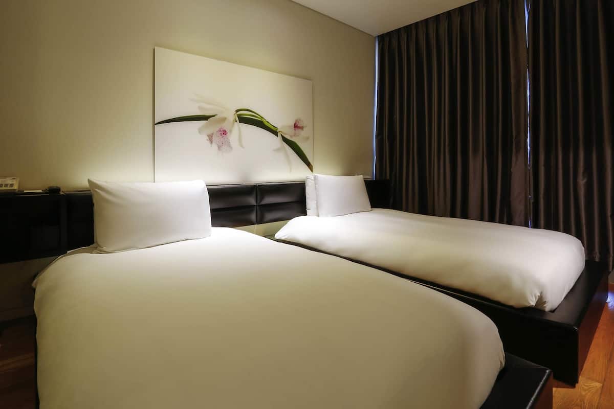 A hotel room with two beds, a dark headboard, a wall-mounted artwork of flowers, and a window with dark curtains.