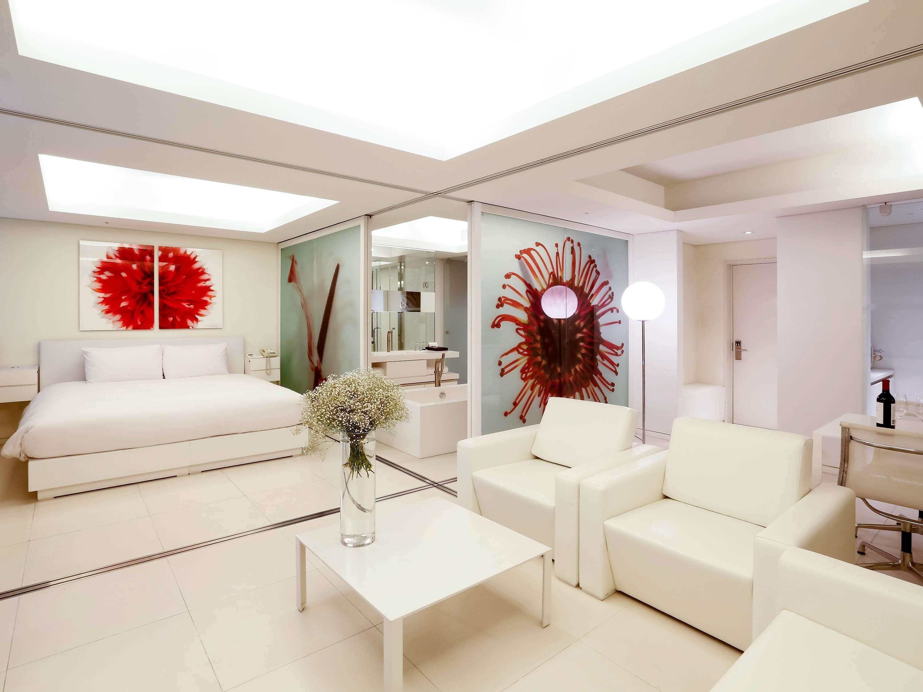 A modern hotel room with a bed, a sofa, a coffee table, a vase with flowers, a large mirror, and a red abstract artwork on the wall.