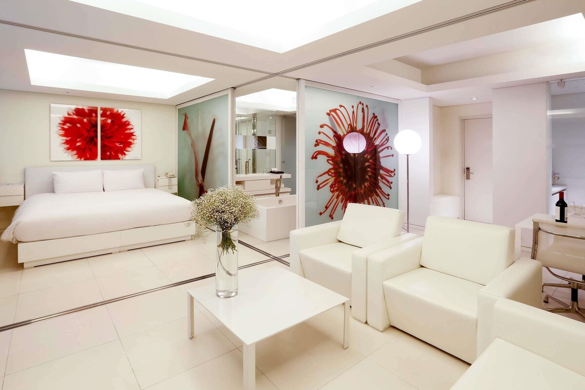 A modern hotel room with a bed, a sofa, a coffee table, a vase with flowers, a large mirror, and a red abstract artwork on the wall.