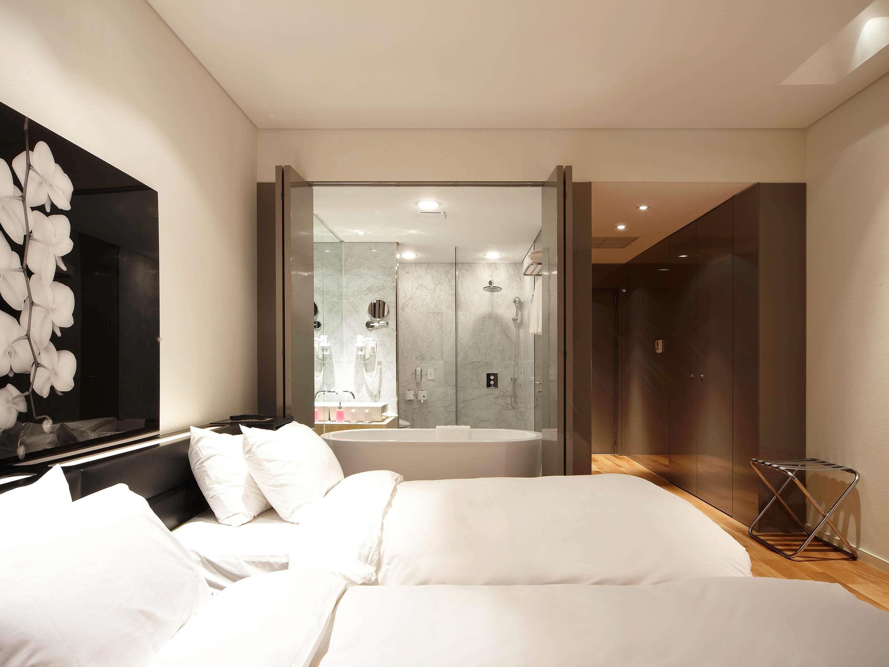 A modern bedroom with a large bed, a bathroom with a glass shower enclosure, and a hallway leading to a dark room.