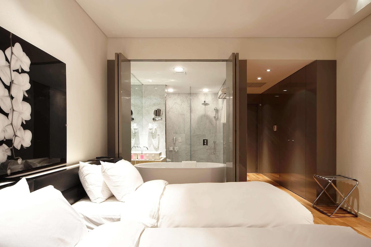A modern bedroom with a large bed, a bathroom with a glass shower enclosure, and a hallway leading to a dark room.
