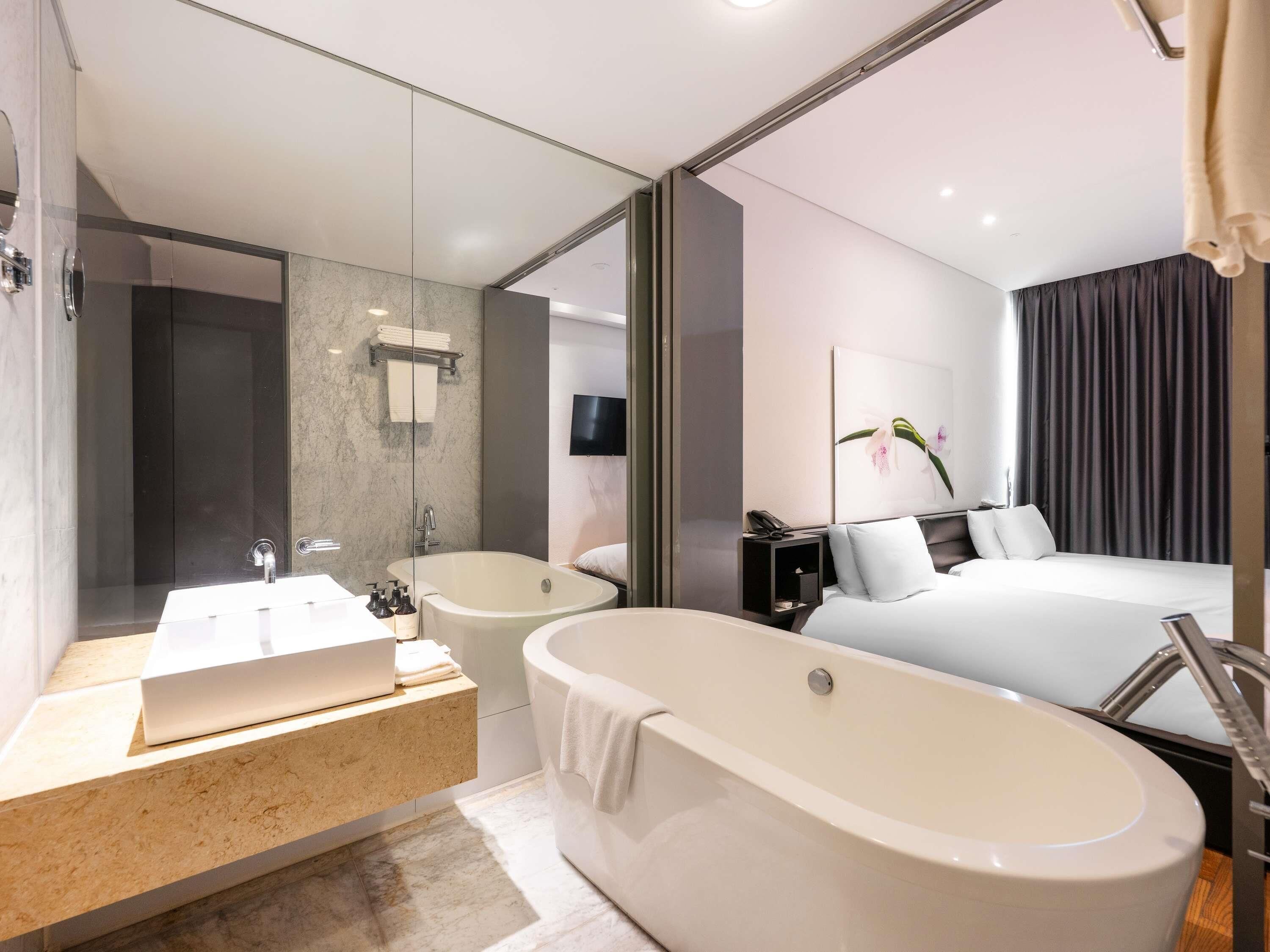 A modern hotel room with a large bathtub, a separate shower area, a vanity with a mirror, and a bed with a view of the room.