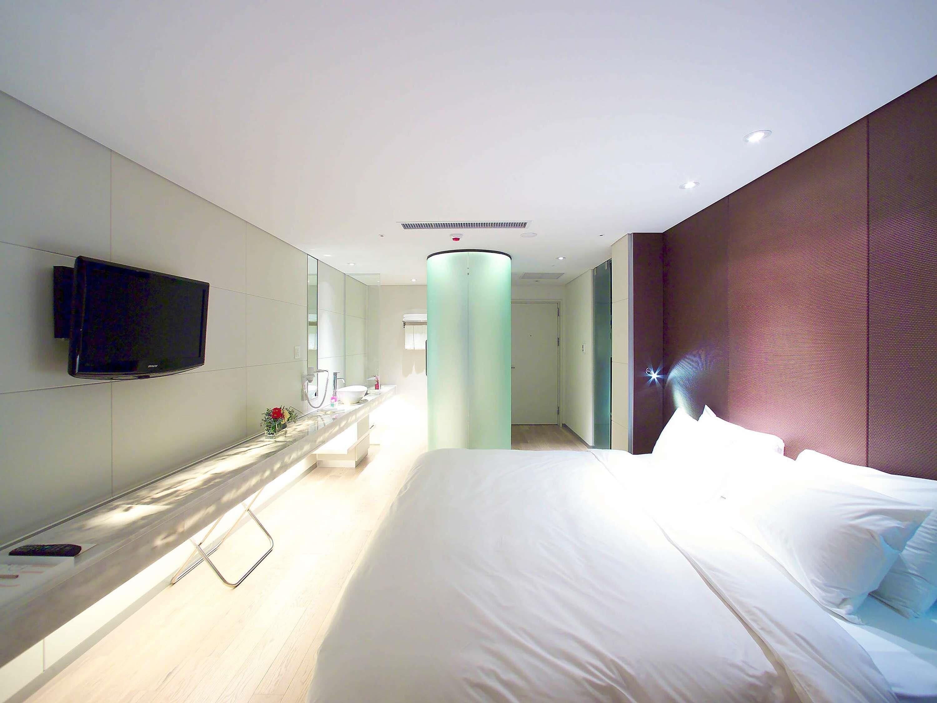 Ein modernes Hotelzimmer mit Bett, einem an der Wand befestigten Flachbildfernseher, einem Badezimmer mit Spiegel und einem Gang zu einem weiteren Zimmer.