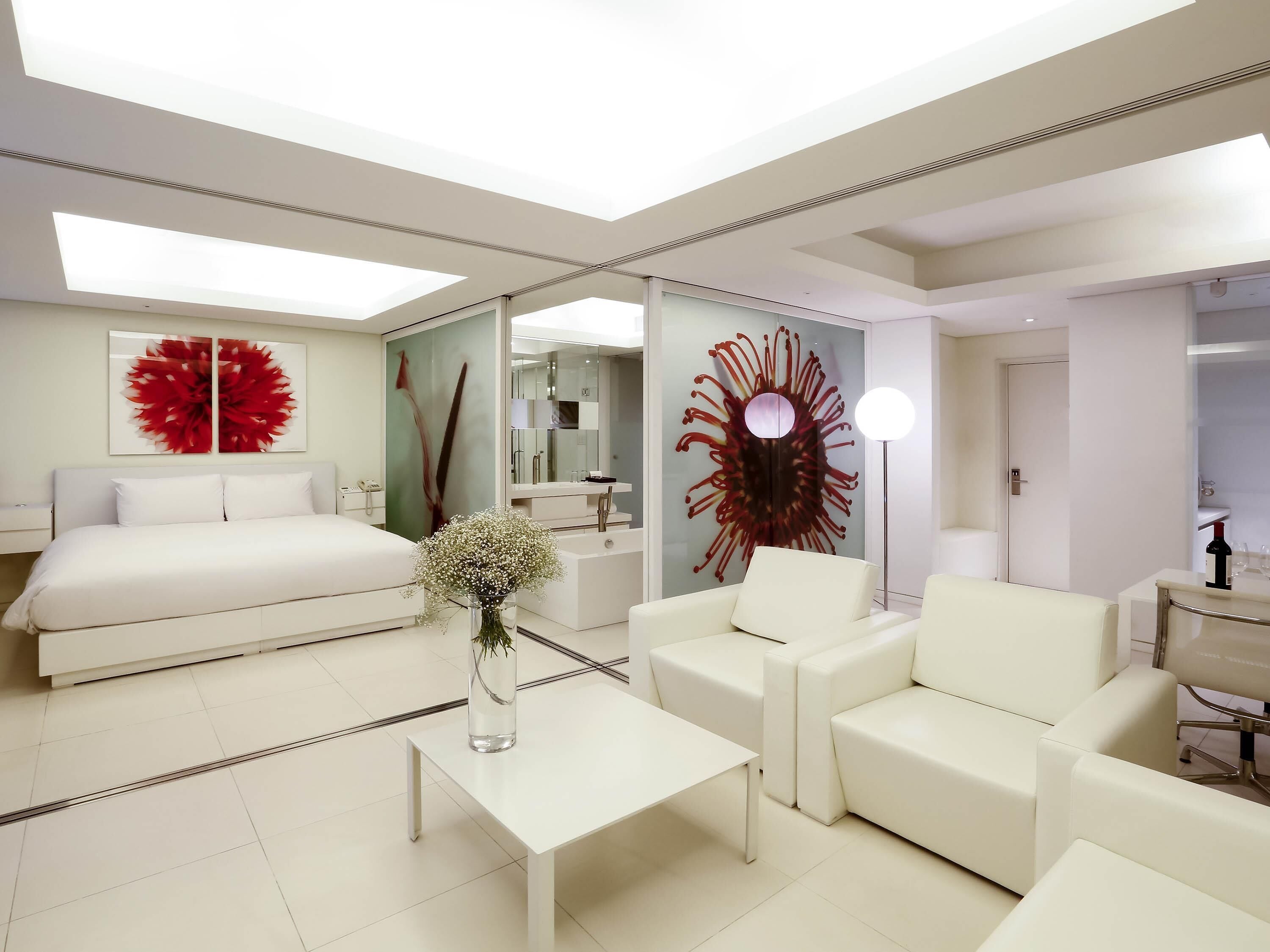 A modern hotel room with a bed, a sitting area with a coffee table, a large mirror, and a decorative wall piece.