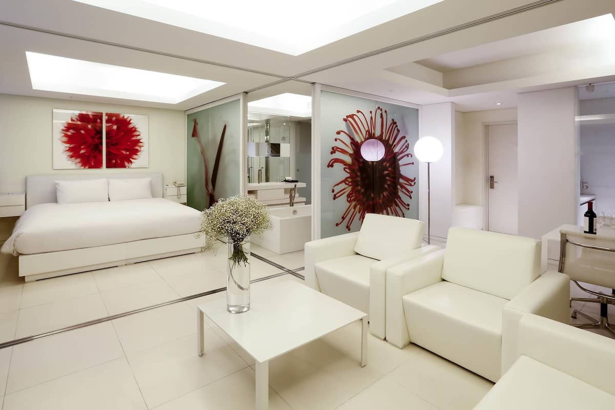 A modern hotel room with a bed, a sitting area with a coffee table, a large mirror, and a decorative wall piece.