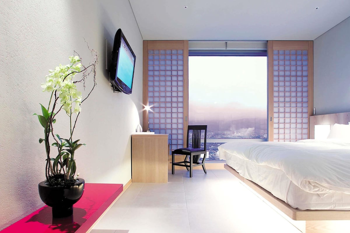 A modern hotel room with a bed, a TV, a chair, and a large window with a city view.