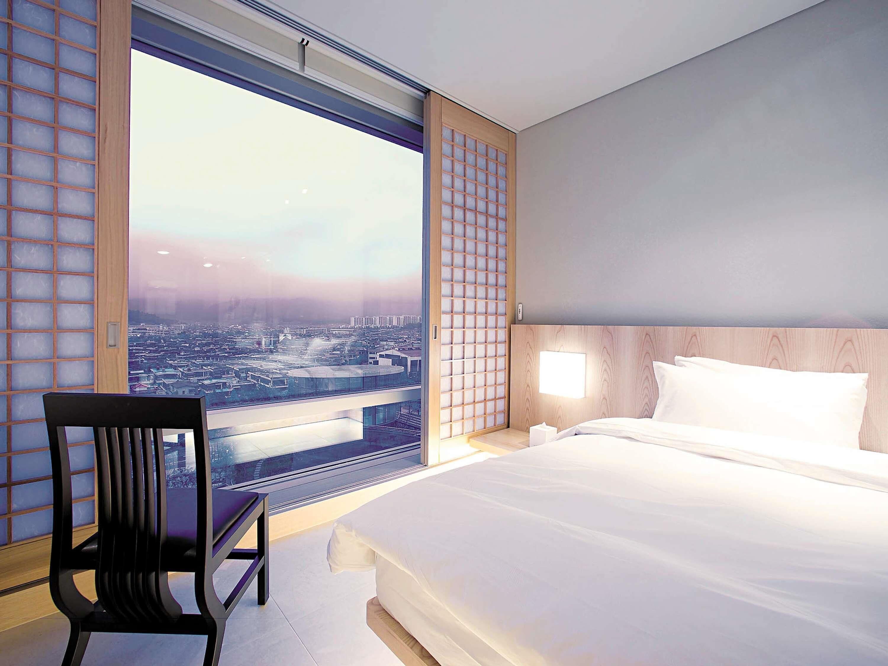 Une chambre d’hôtel avec un grand lit, une chaise et une vue sur le paysage urbain, visible à travers une grande fenêtre.