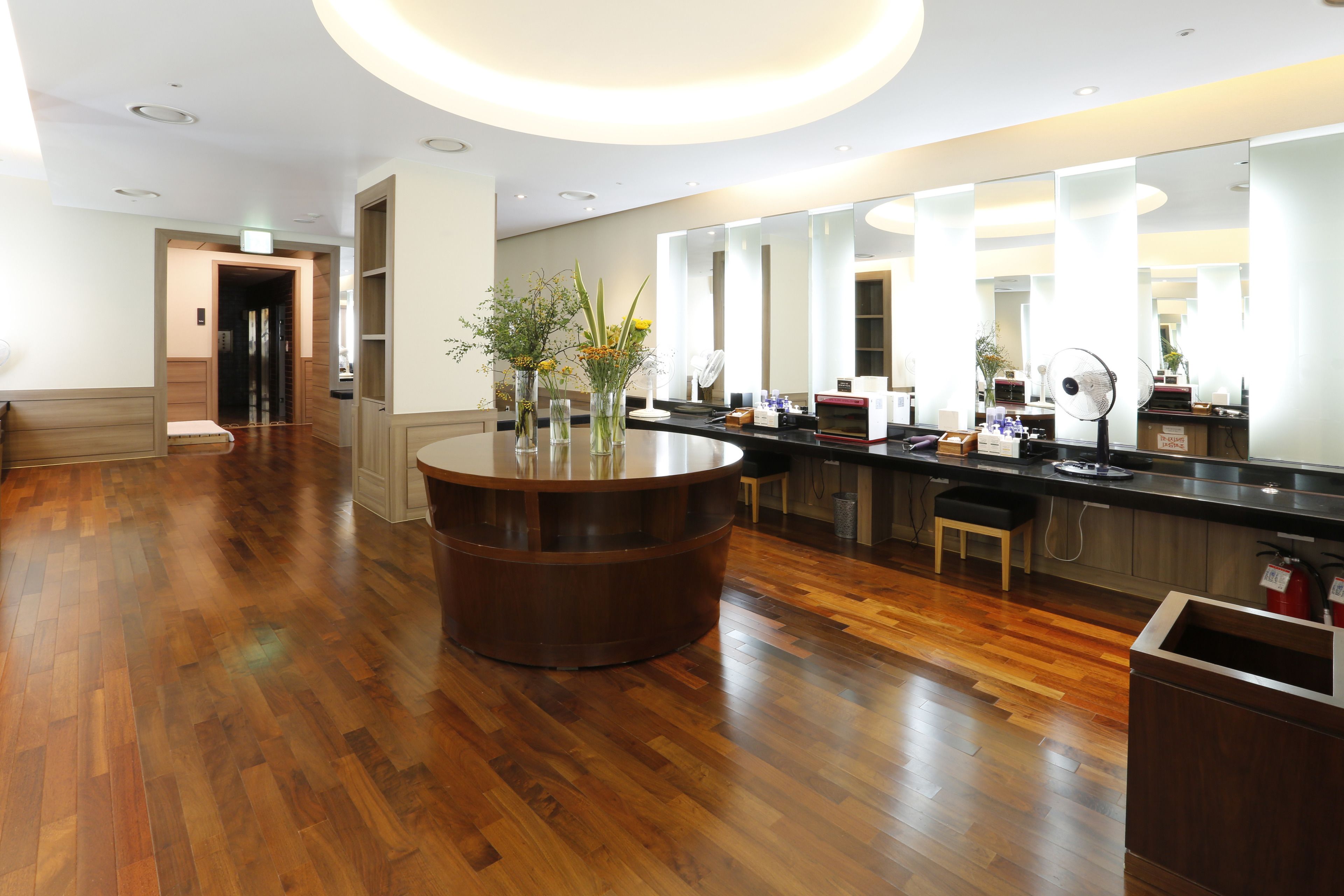 A modern hotel lobby with a wooden floor, a central round table with vases, a long counter with a mirror, and a fire extinguisher.