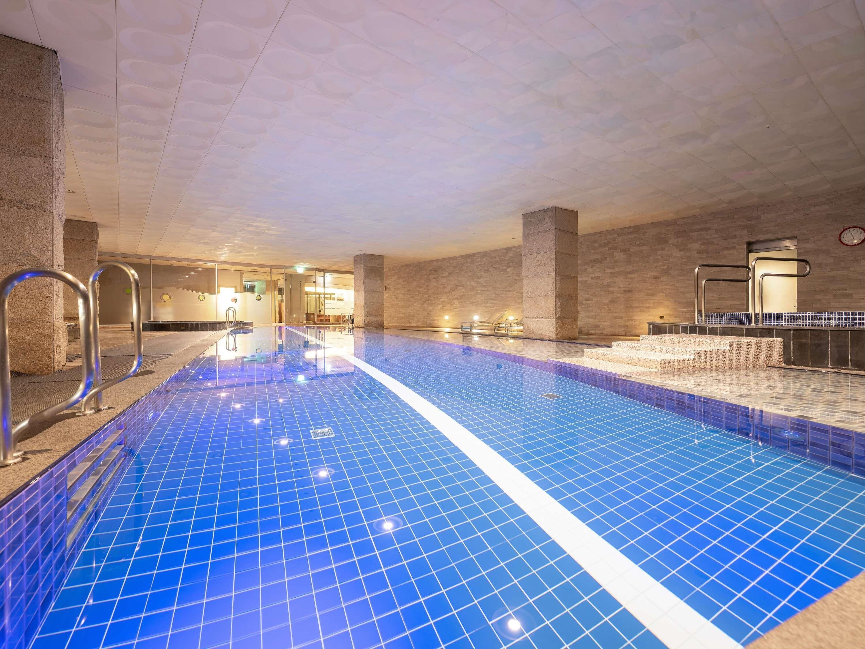 An indoor swimming pool with clear blue tiles, metal handrails, and a tiled wall.