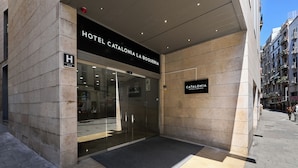 La entrada al HOTEL CATALONIA LA BOQUERIA, con fachada de vidrio y un felpudo negro en la entrada.