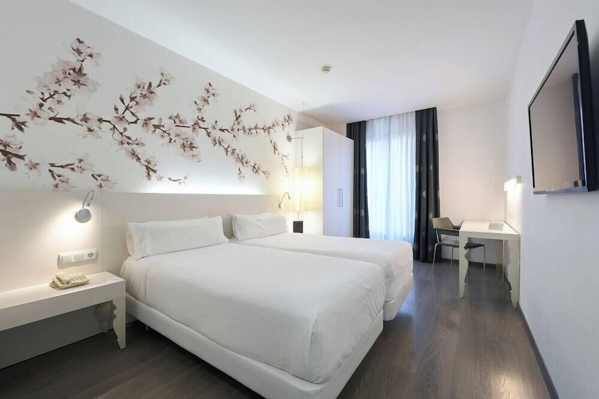 A hotel room with two beds, a desk, a chair, a flat-screen TV, and a wall-mounted lamp.
