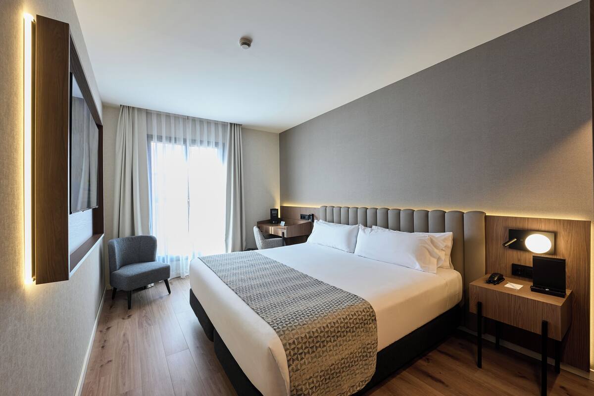 A hotel room with a large bed, bedside tables, a chair, a desk, and a TV.
