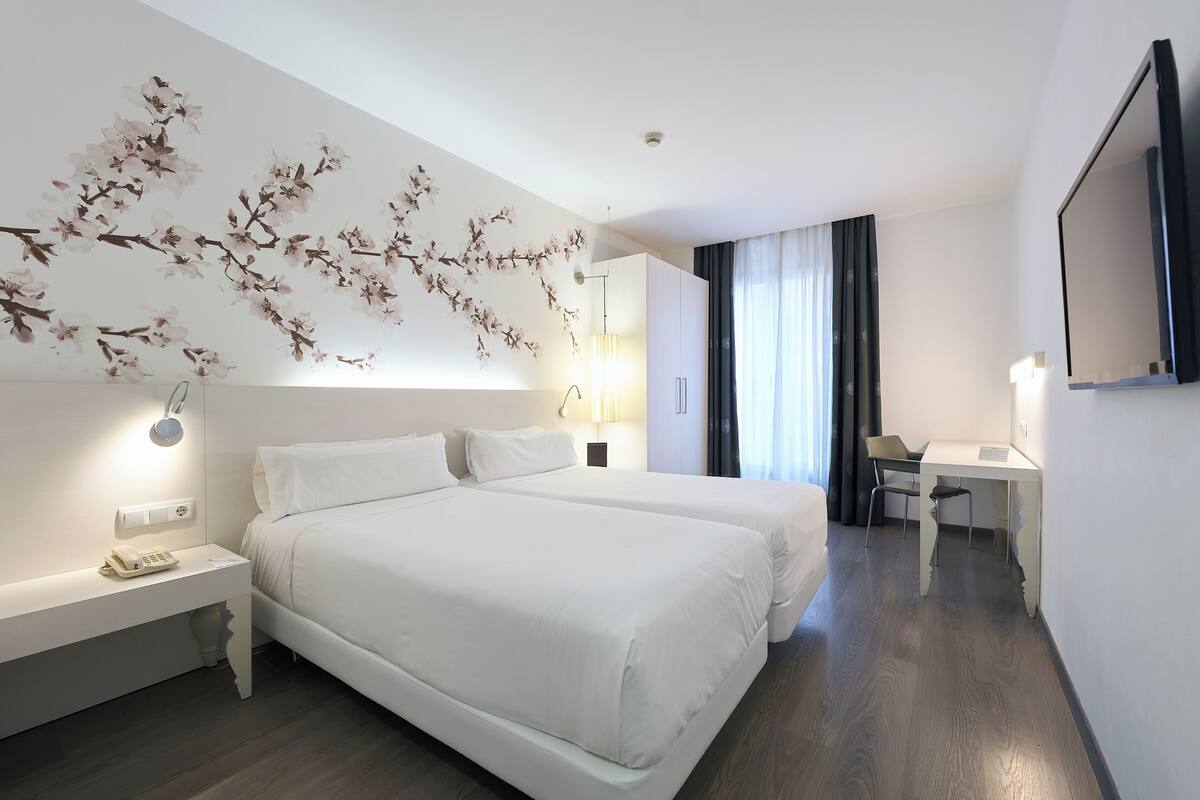 A hotel room with two beds, a desk, a chair, a lamp, and a wall-mounted TV.