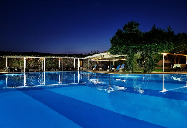 Una piscina con agua azul cristalina, rodeada de sillas de descanso y una zona cubierta con mesas y sillas.