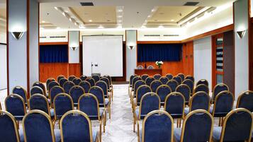 Uma sala de conferências com fileiras de cadeiras azuis, um pódio e uma tela de projeção.
