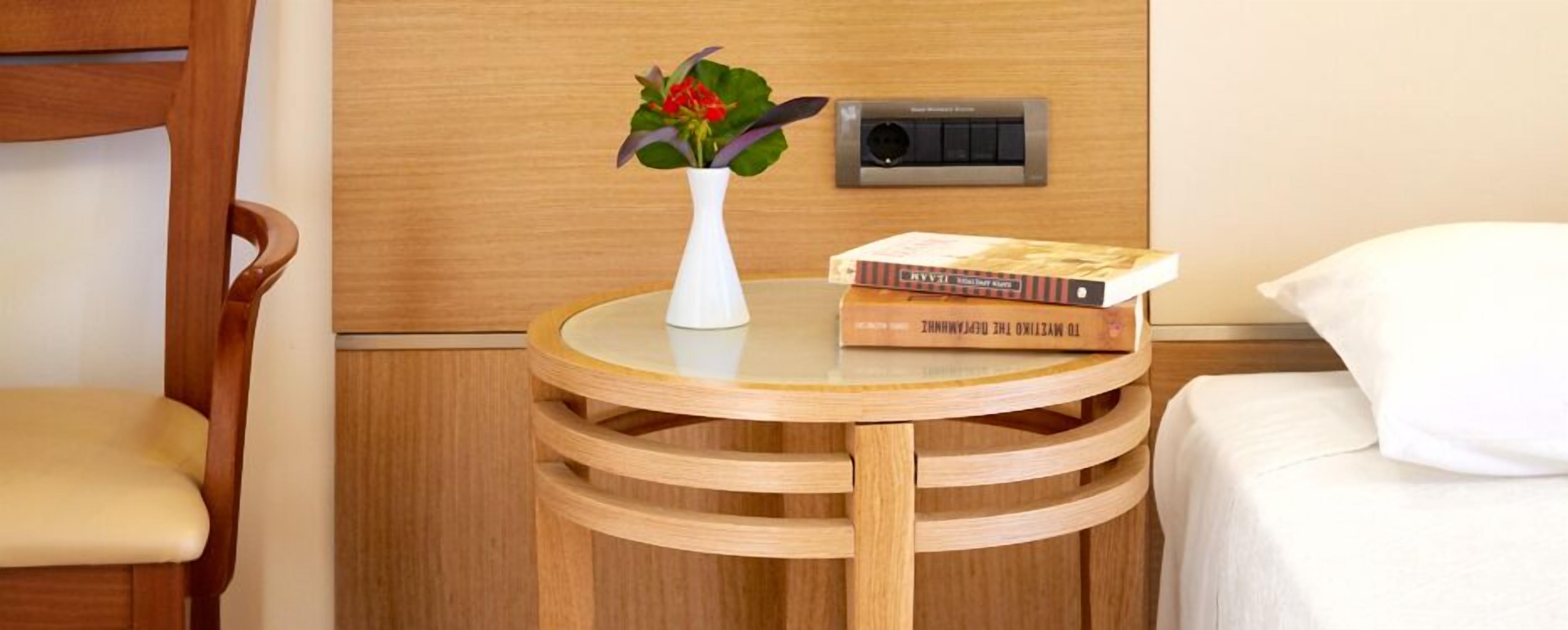 A wooden chair, a small round table with a white vase and a book, and a bed with a white pillow.