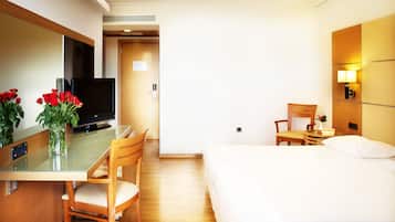 Habitación de hotel con cama, escritorio con televisor, silla, mesita de noche y un jarrón con flores.