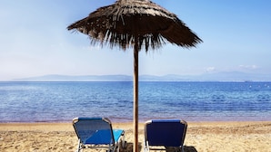Une scène de plage avec deux chaises de plage bleues sous un parasol en chaume, une plage de sable et une mer calme avec des montagnes à l’horizon.