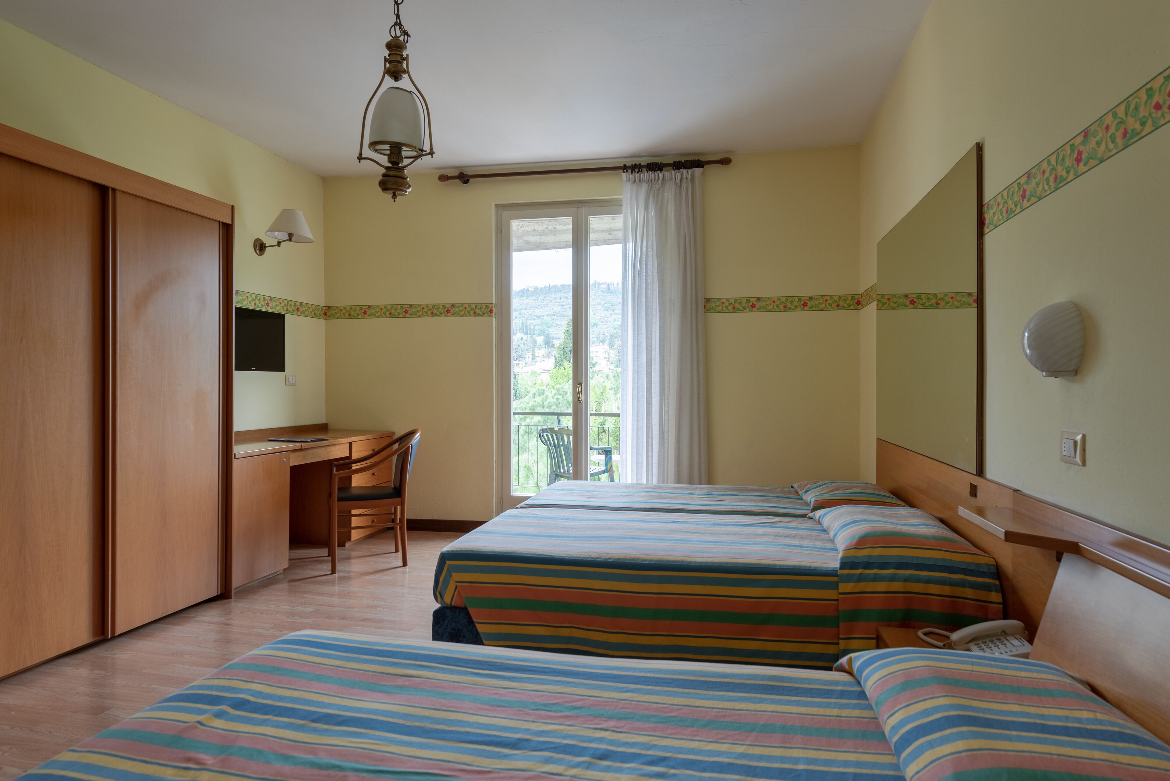 A hotel room with two beds, a desk, a chair, and a balcony with a view.