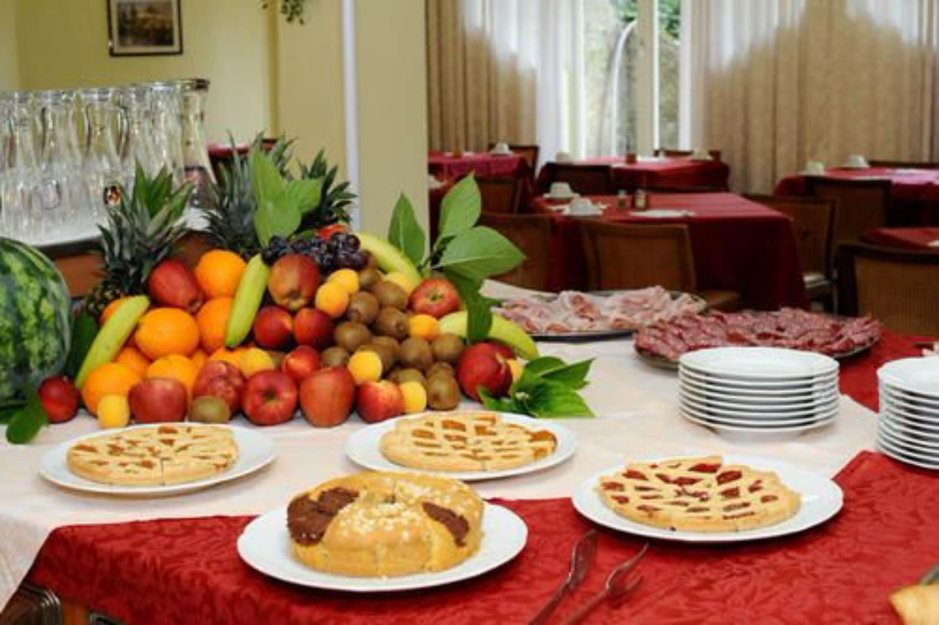 A dining area with tables set for a meal, fruit and dessert displayed on the table, and a stack of plates.