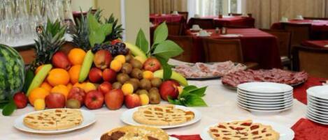 A dining area with tables set for a meal, fruit and dessert displayed on the table, and a stack of plates.
