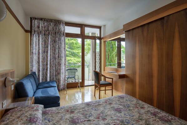 A room with a bed, a desk, a chair, and a sofa. There is a window with curtains, and a view of greenery outside.