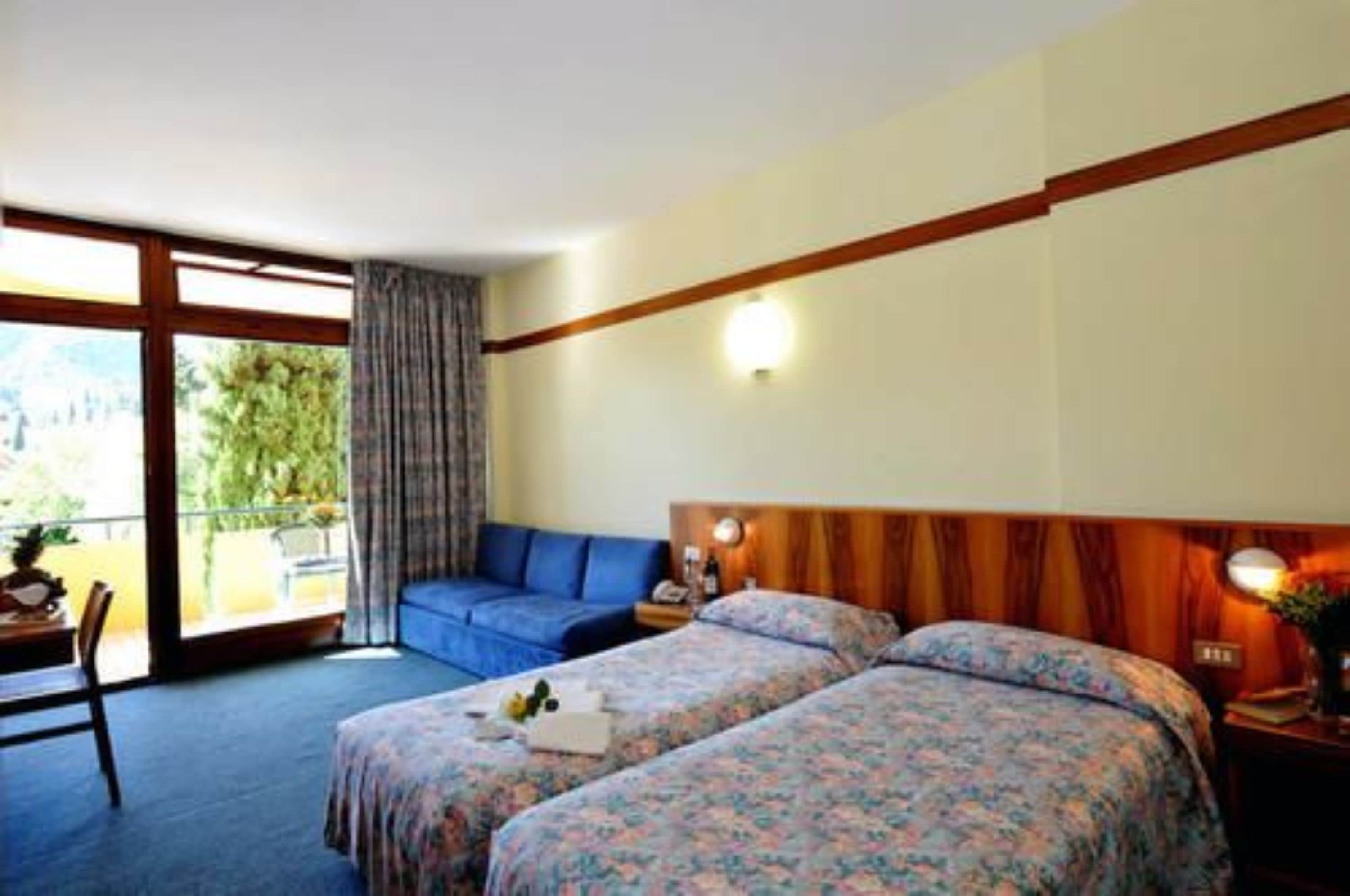 Ein Hotelzimmer mit zwei Betten, einem Sofa, einem Schreibtisch und einem Balkon mit Blick auf Grünflächen.
