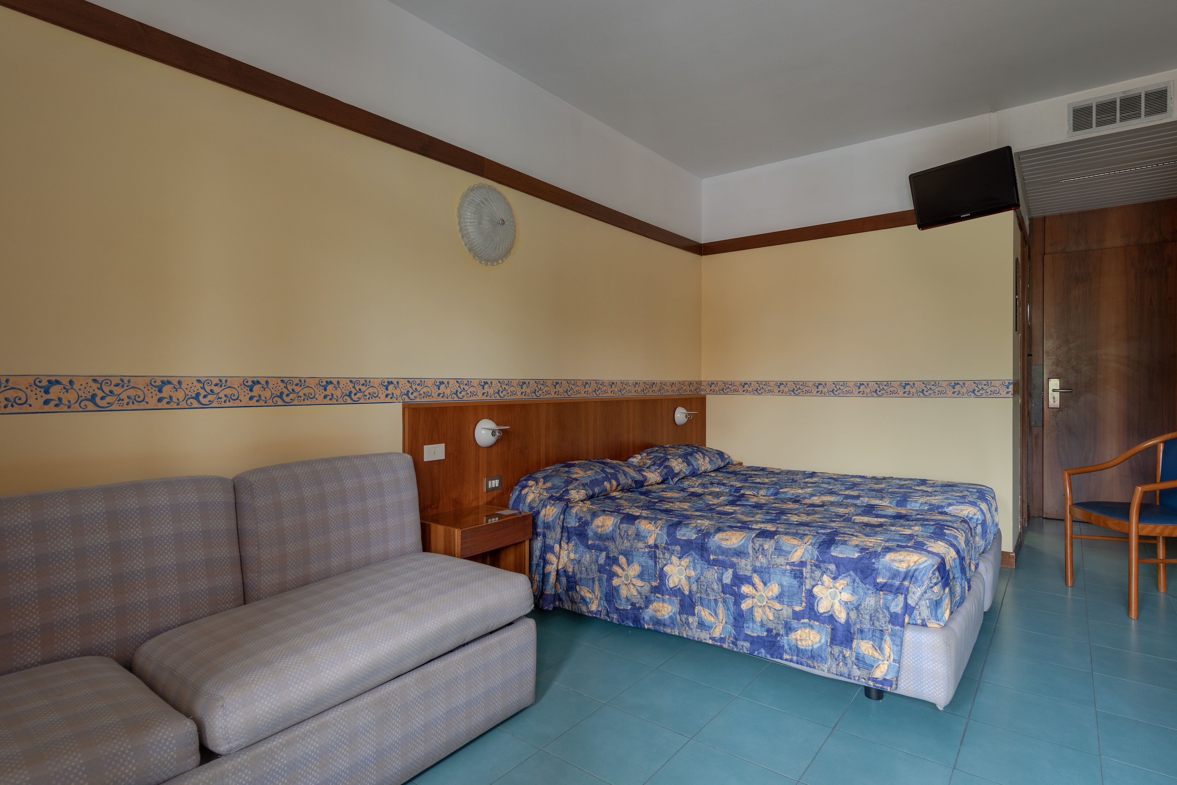Habitació amb llit, sofà, capçalera de fusta, rellotge de paret i televisió penjada a la paret.