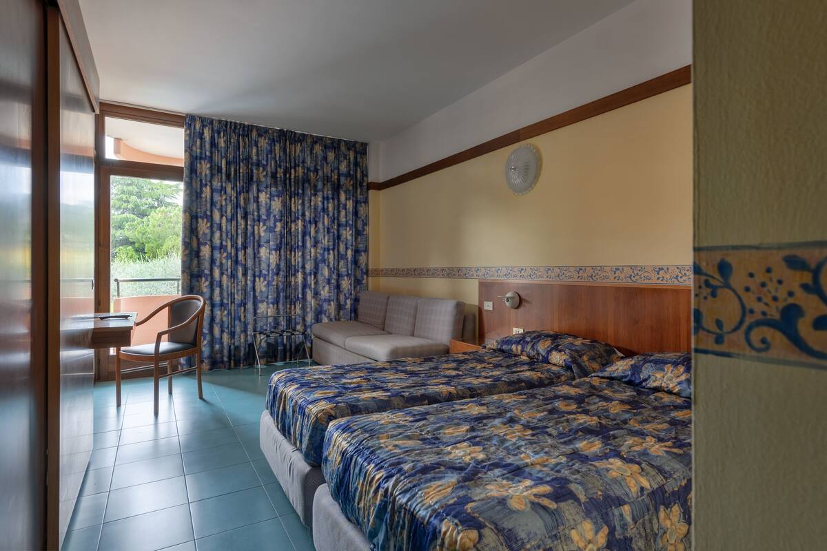 A hotel room with two beds, a desk, a chair, and a view of greenery outside.