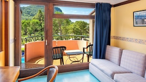 A balcony with a table and chairs, a sofa, and a view of a pool and trees. - Hotel Royal (Garda)