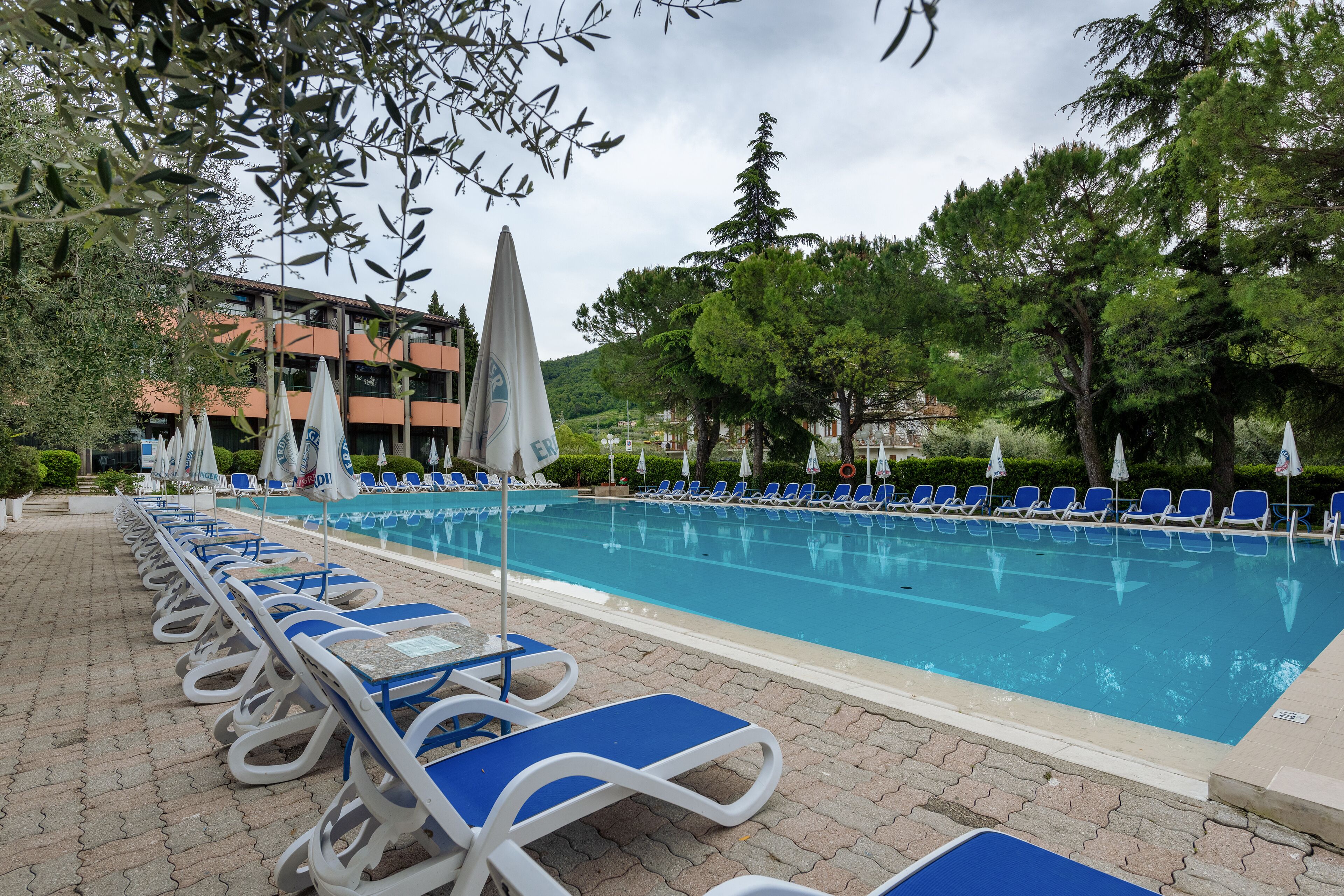 A pool area with blue lounge chairs and umbrellas, surrounded by trees and buildings.