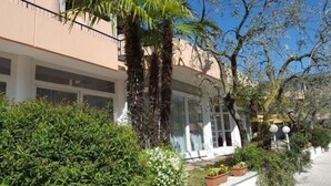 A building with a beige facade, large windows, and a garden area with potted plants and a palm tree. - Hotel Royal (Garda)